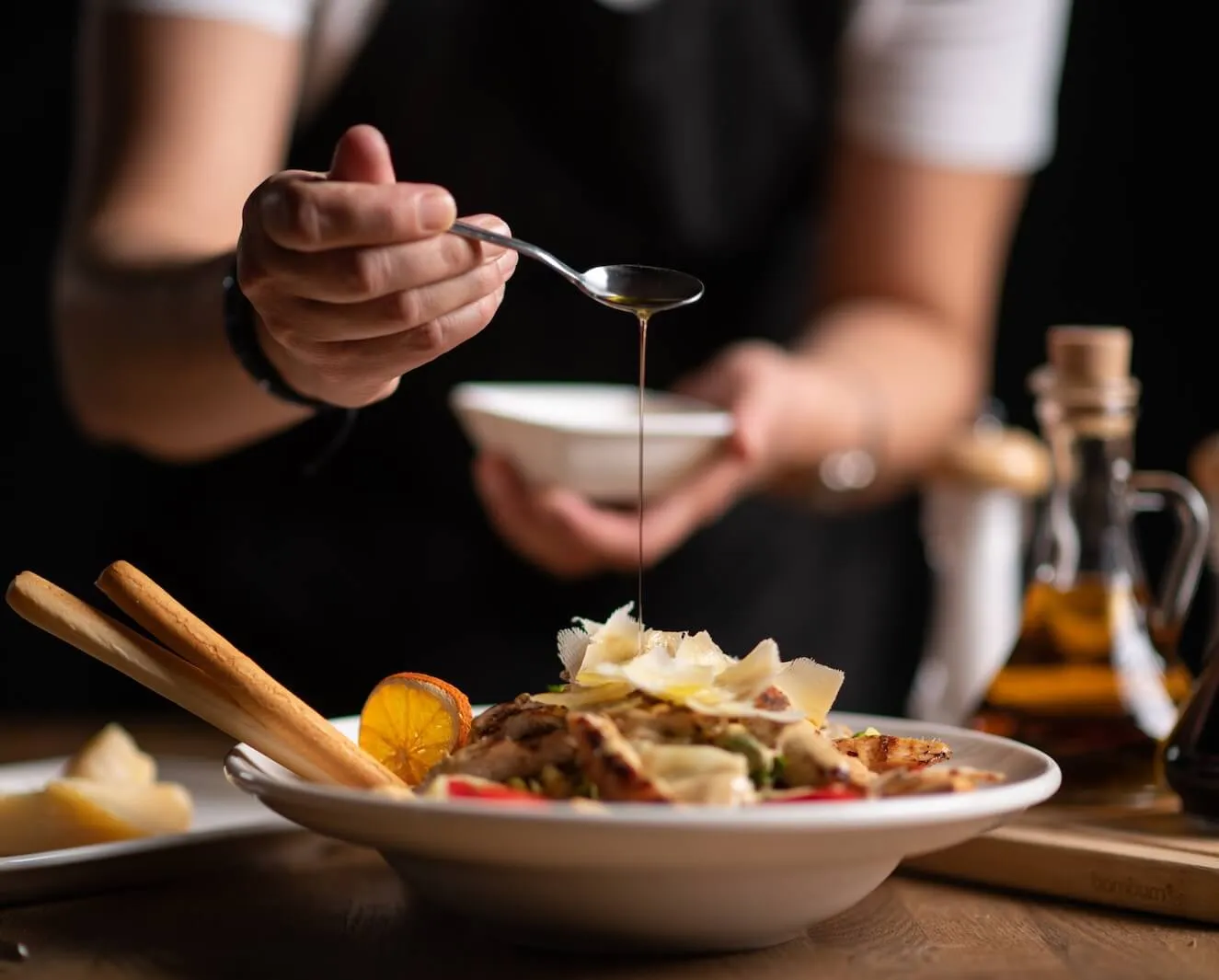 A female chef pouring sauce from the spoon on meal on a black background.