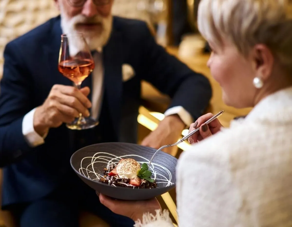 Close image of hand with glass of champagne, sweet dessert on grey dish. Bearded man in tuxedo drinking