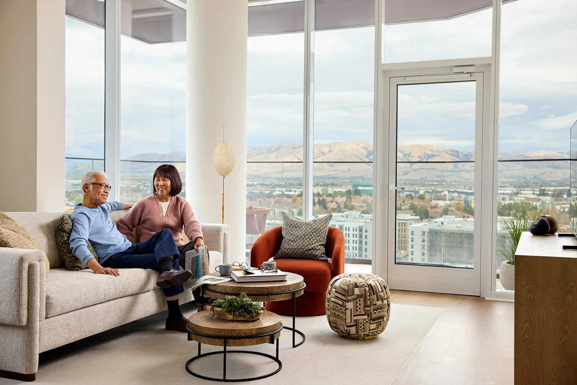 A senior couple relaxing together on a cream sofa in a bright, modern apartment living room at Ellore Senior Living, smiling and conversing surrounded by nesting coffee tables, a rust accent chair, and wraparound floor-to-ceiling windows with panoramic mountain and city views.