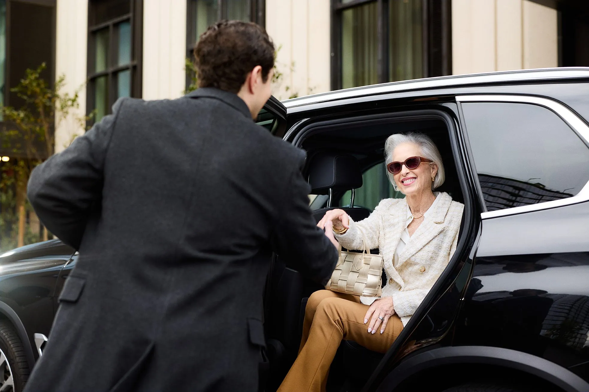 A well-dressed senior woman smiling as a concierge assists her out of a black luxury vehicle outside Ellore Senior Living, conveying personalized transportation and white-glove service.