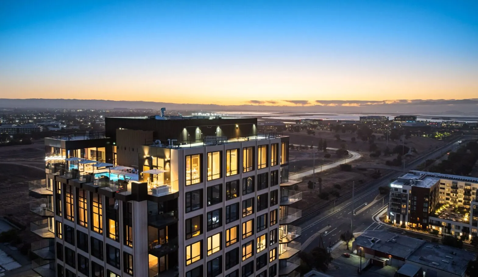 Aerial dusk view of the Ellore Senior Living tower with warmly lit apartments, an illuminated rooftop pool and terrace, and a sweeping panorama of the surrounding cityscape, bay, and mountains beneath a vivid orange and blue sunset sky.