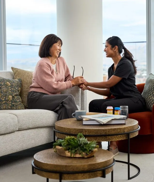 A smiling senior resident and a care staff member hold hands warmly during a conversation in a bright, modern living room at Ellore Senior Living, with mountain views visible through floor-to-ceiling windows in the background.