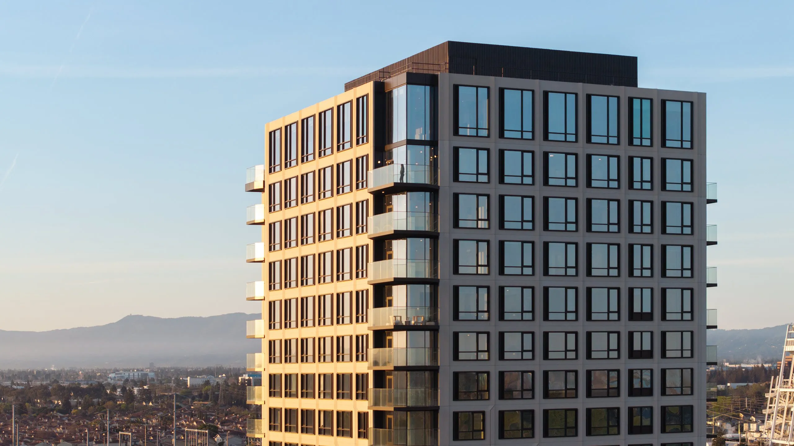 Aerial exterior view of the Ellore Senior Living exterior at golden hour, featuring a modern facade with large windows, glass-railed balconies, and a dark rooftop penthouse level, set against a cityscape and mountain backdrop.