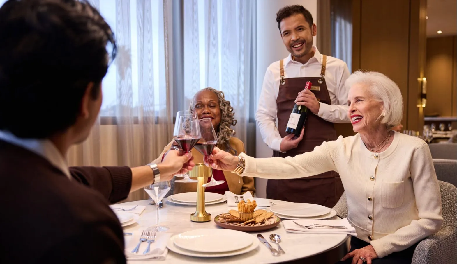 Residents raise a toast over a beautifully set table as an attentive server presents a bottle of red wine, capturing the fine dining experience and warm social atmosphere at Ellore Senior Living.