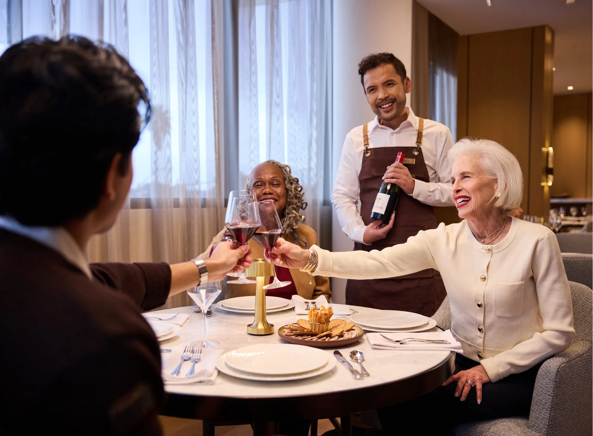 Residents raise a toast over a beautifully set table as an attentive server presents a bottle of red wine, capturing the fine dining experience and warm social atmosphere at Ellore Senior Living.