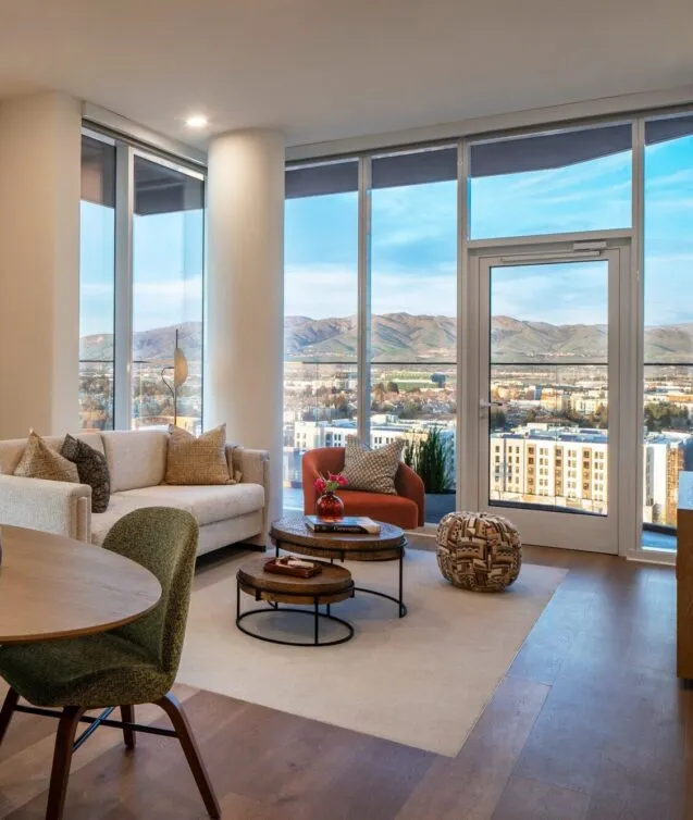Bright, modern living room in a spacious senior living apartment at Ellore, featuring floor-to-ceiling windows with panoramic mountain and city views, a cream sofa, rust-colored accent chair, nesting coffee tables, and warm hardwood floors.