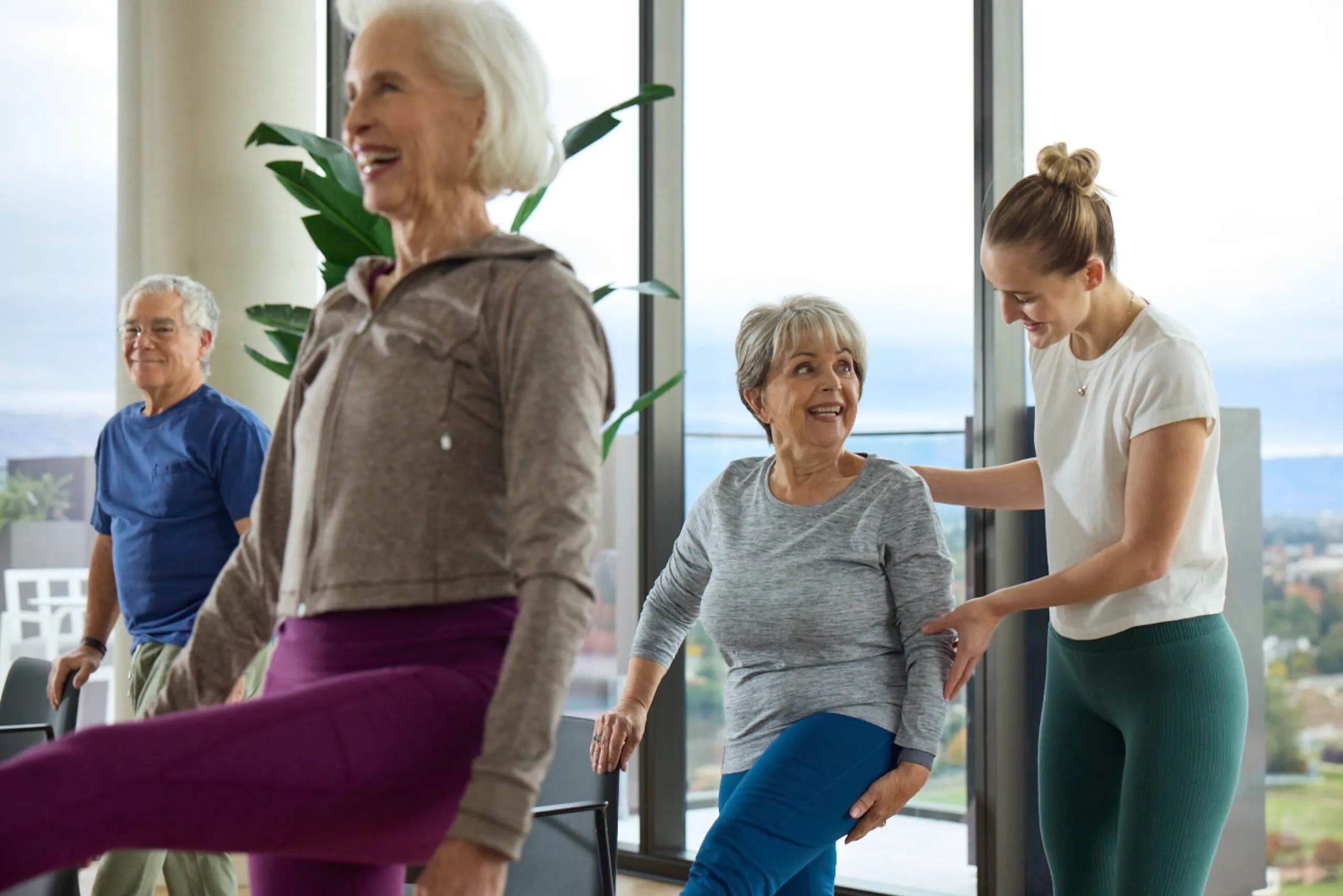 Three senior residents participate in a group exercise class while a fitness instructor provides hands-on guidance to one resident, all smiling in a bright, windowed fitness studio with city views at Ellore Senior Living.