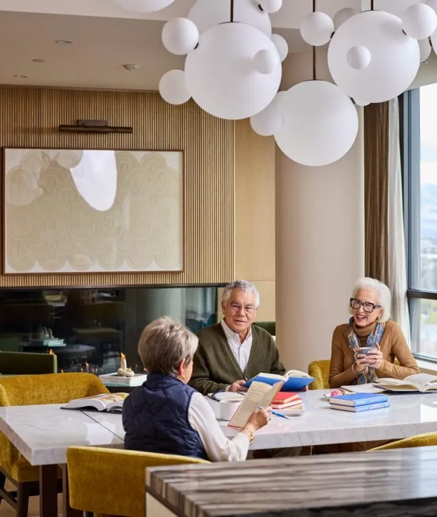 Three senior residents gathered around a marble table for a book club, laughing and reading together in an elegant common room at Ellore Senior Living, featuring a sculptural bubble chandelier, wood-slat fireplace wall, gold velvet chairs, and mountain views through floor-to-ceiling windows.