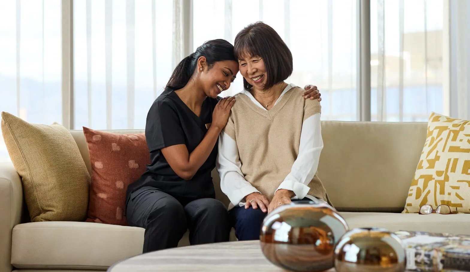 A care staff member and a senior resident share a warm, laughing embrace seated side by side on a sofa in a bright, elegantly furnished apartment living room at Ellore Senior Living.