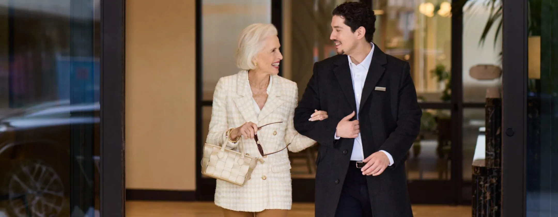 A smiling senior resident walks arm-in-arm with a uniformed concierge through the entrance of Ellore Senior Living, both laughing and conversing in a warm lobby setting.