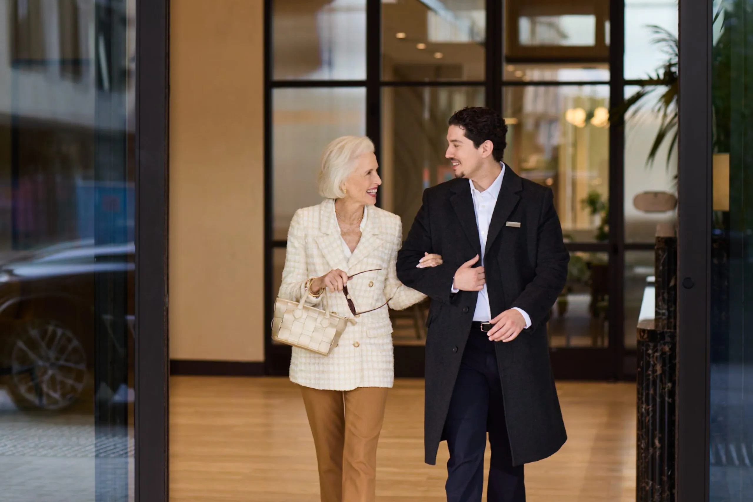 A smiling senior resident walks arm-in-arm with a uniformed concierge through the entrance of Ellore Senior Living, both laughing and conversing in a warm lobby setting.