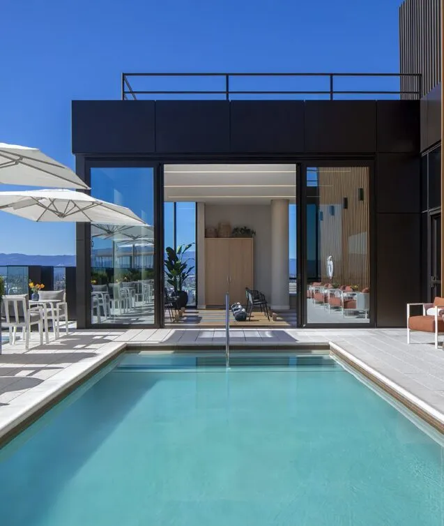 Rooftop pool and terrace at Ellore Senior Living featuring a rectangular lap pool, outdoor seating with rust-colored chairs and white umbrellas, warm wood-slat wall cladding, and panoramic mountain views under a bright blue sky.